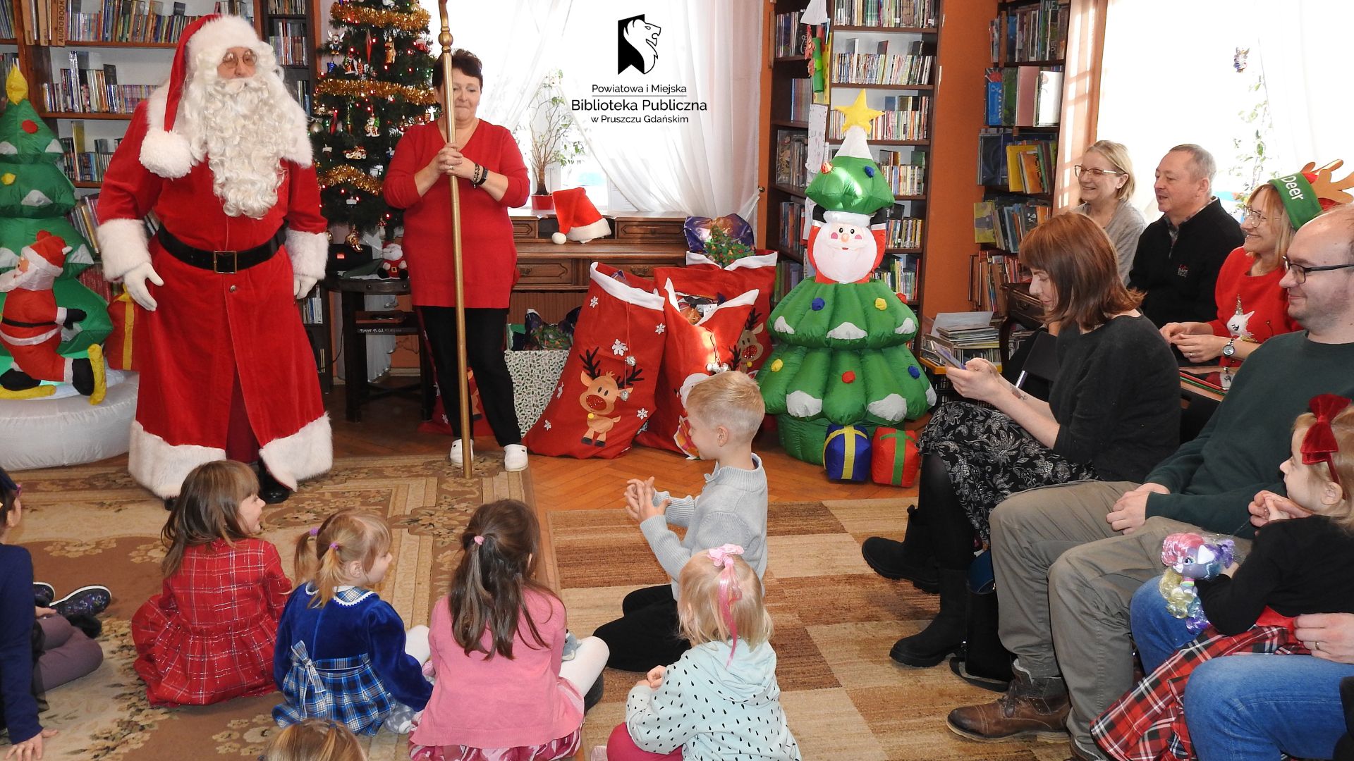 Na dywanie w bibliotece siedzą dzieci i patrzą w stronę mężczyzny w stroju Mikołaja. Obok mężczyzny stoi kobieta w czerwonym swetrze i trzyma laskę Mikołaja. Po prawej i lewej stronie stoją dmuchane ozdoby świąteczne: choinki, bałwany.