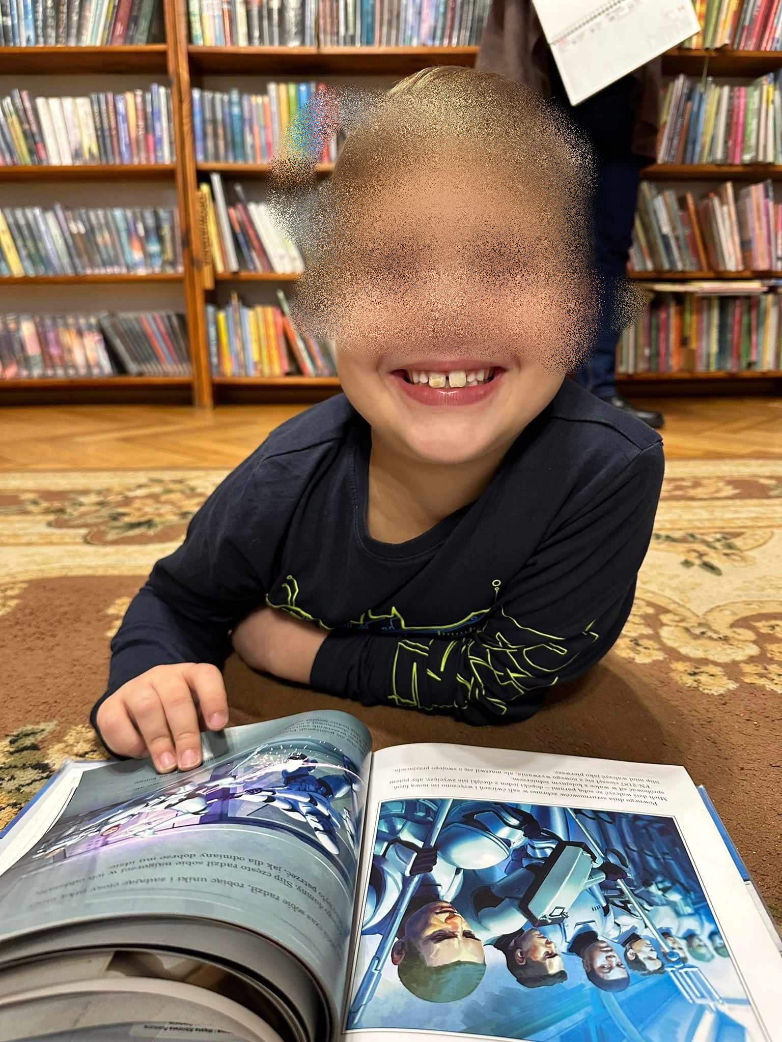 Na dywanie, na brzuchu, leży uśmiechnięty chłopiec. Przed chłopcem leży otwarta książka.
