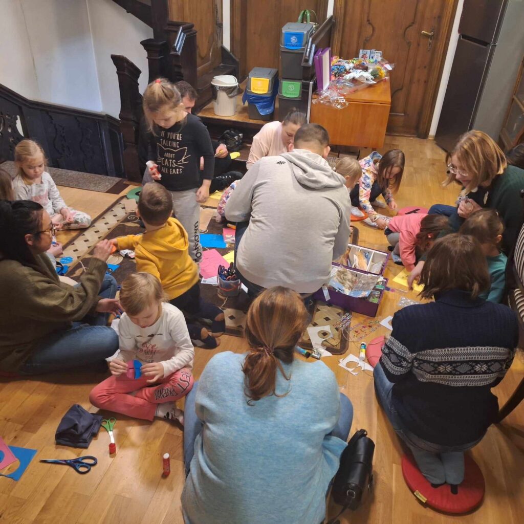 Na kolorowych poduszkach siedzą dzieci i rodzice. Wszyscy robią bombki z papieru metodą origami. Obok leżą nożyczki, kleje, kolorowe cekiny, papier.