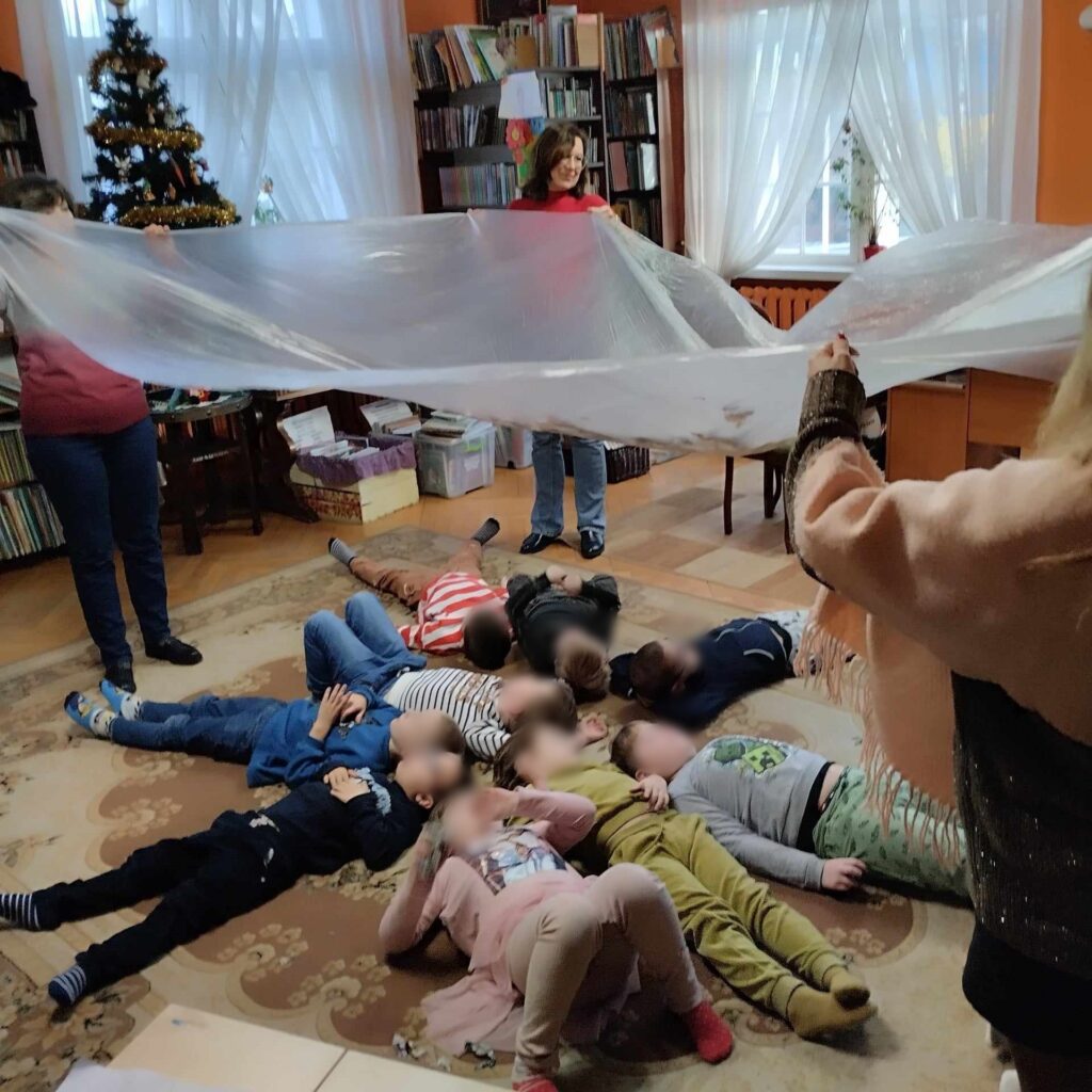 Na dywanie leżą dzieci. Na ich głowami bibliotekarka i nauczycielki machają przezroczystą folią.