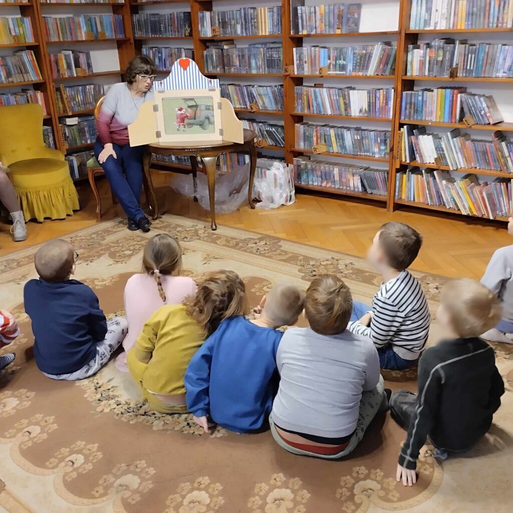 Dzieci siedzą na podłodze i patrzą w stronę teatrzyku kamishibai, który stoi na stoliku. Po lewej siedzi bibliotekarka i czyta. W tle regały z książkami.