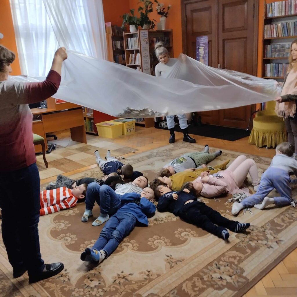 Na dywanie leżą dzieci. Na ich głowami bibliotekarka i nauczycielki machają przezroczystą folią, na której leżą skrawki gazety.