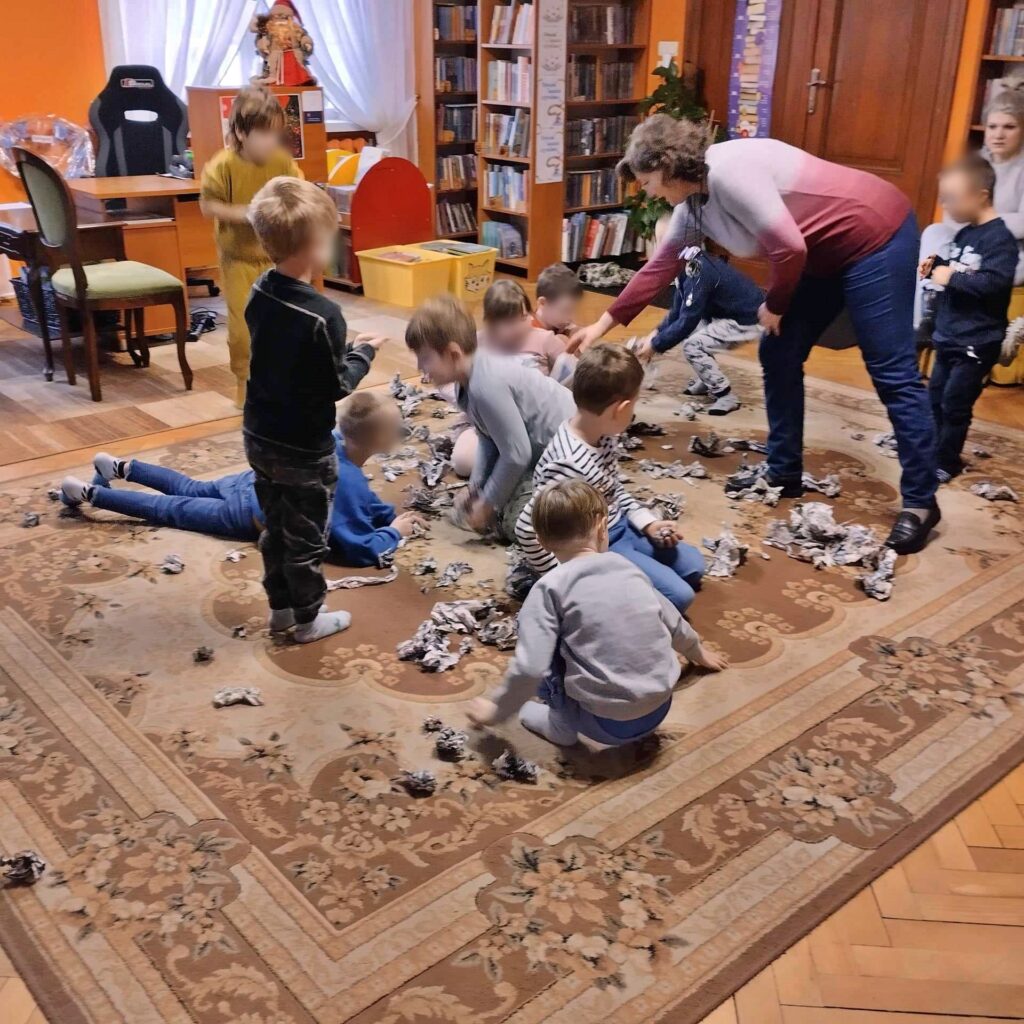 Dzieci siedzą i stoją na dywanie. Między nimi leżą skrawki gazet. Obok dzieci stoi bibliotekarka i wskazuje palec na skrawki papieru.