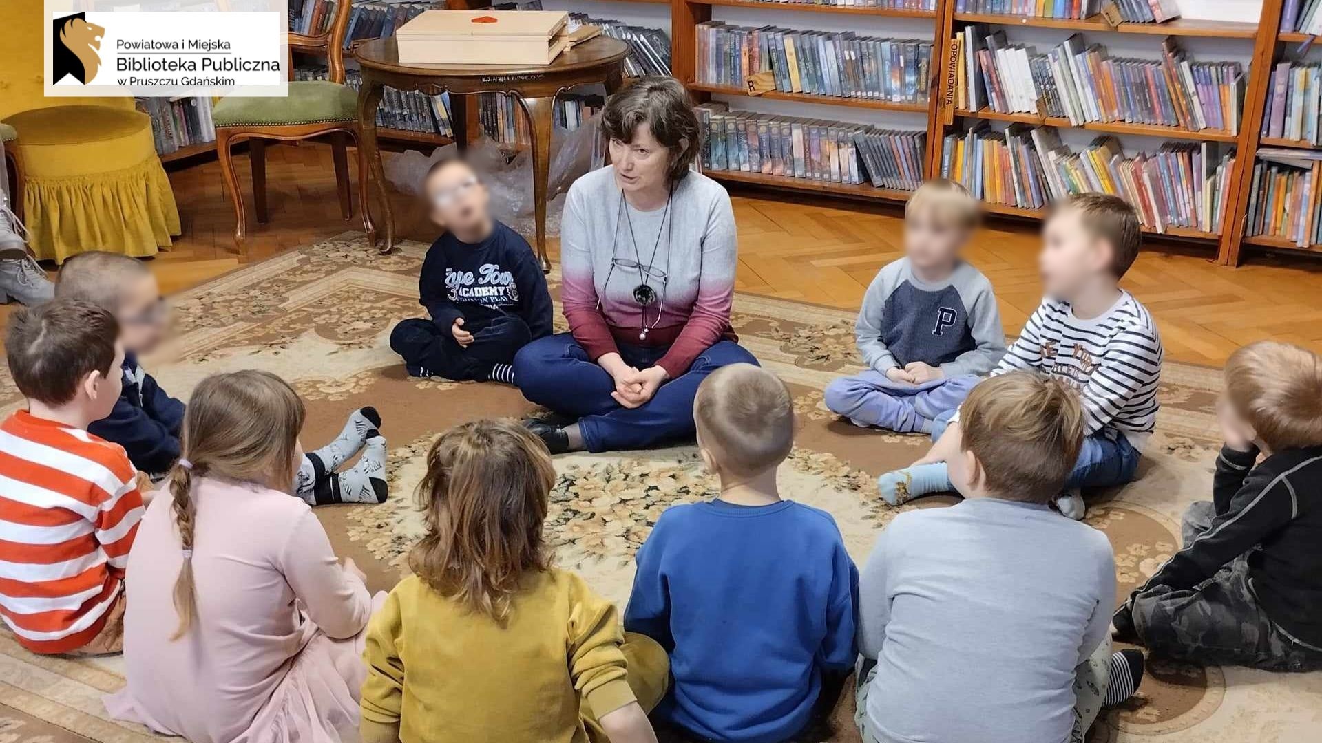 Grupa dzieci siedzi w okręgu na dywanie. Między nimi siedzi bibliotekarka. W tle regały z książkami oraz stolik, na którym leży drewniany teatrzyk.