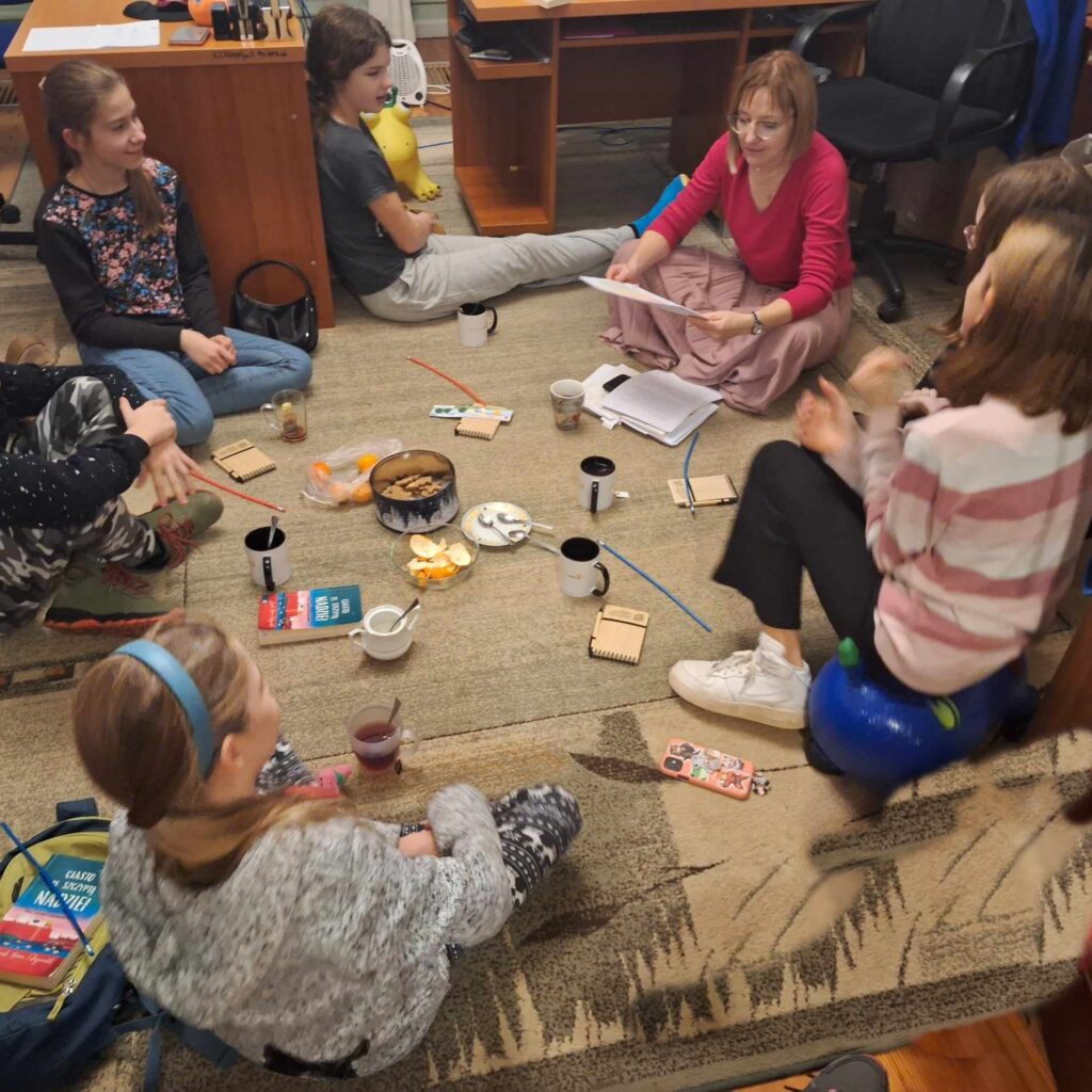 Na dywanie siedzi: 5 dziewczynek, 1 chłopiec. Wszyscy patrzą w stronę moderatorki klubu książki, która czyta z kartki. Pośrodku stoją ciastka w pudełku, kubki z napojem oraz leżą książki.