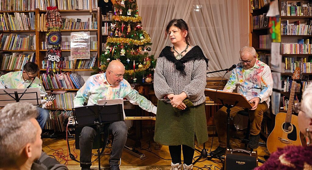 3 mężczyzn z zespołu Bóbr i Kos siedzi na krzesłach z gitarami. Z przodu, przed zespołem, stoi Elena Ulyanova prezentuje muzyków przez publicznością.