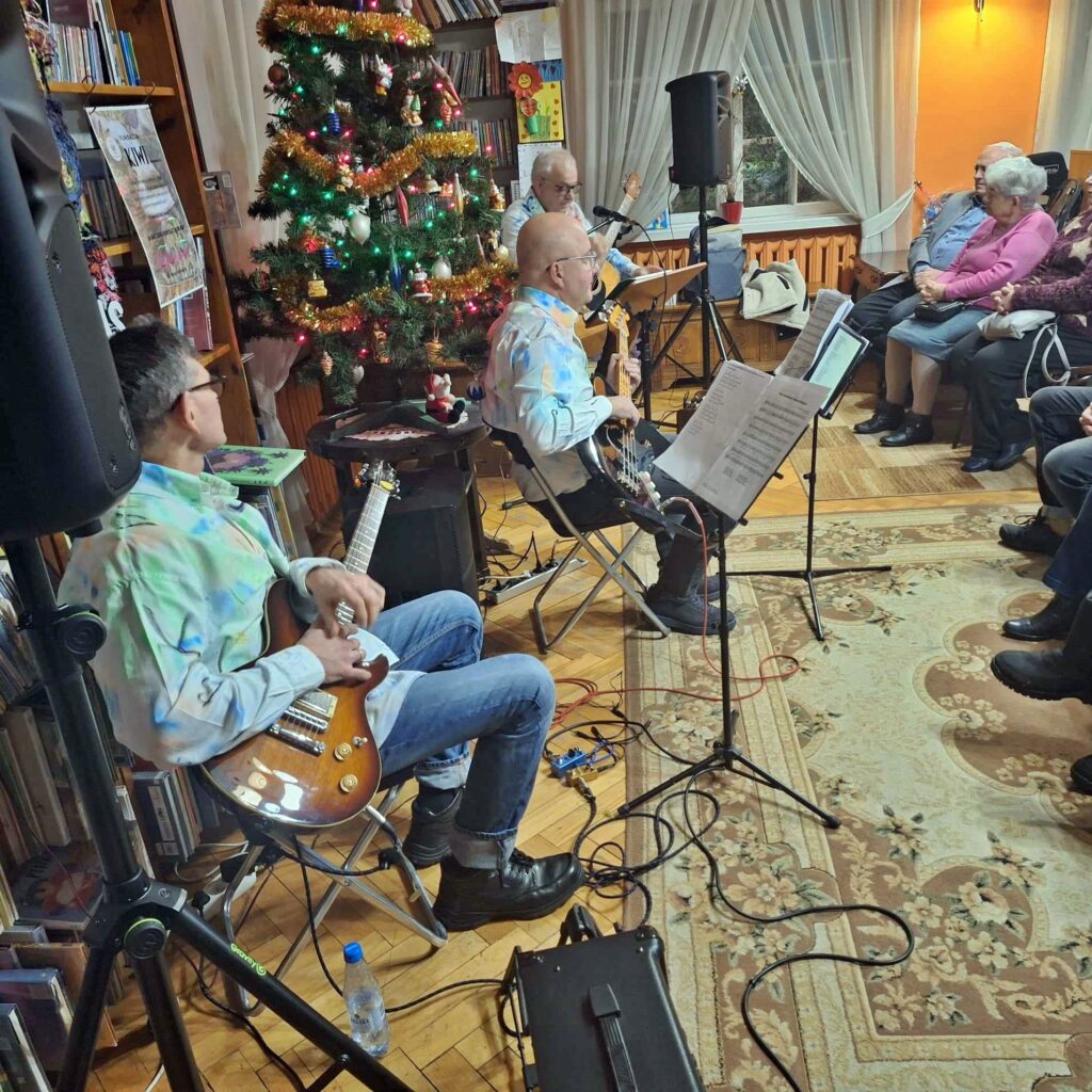 3 mężczyzn z zespołu Bóbr i Kos siedzi na krzesłach z gitarami. Jeden z nich śpiewa. Przed nimi na krzesłach siedzi publiczność.