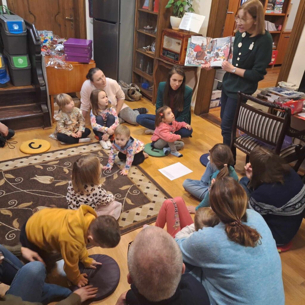 Dzieci oraz rodzice siedzą na kolorowych poduszkach i patrzą w stronę bibliotekarki. Bibliotekarka stoi i czyta książkę Charlotte i Adama Guillain pt. Ile jeszcz do gwiazdki. Pokazuje ilustracje.