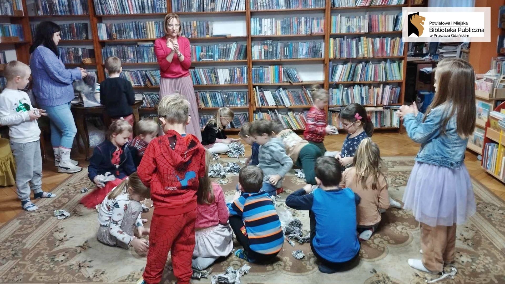 Na dywanie siedzą dzieci wśród papierowych kulek. Przed nimi stoi bibliotekarka i coś mówi. W tle regały z książkami.