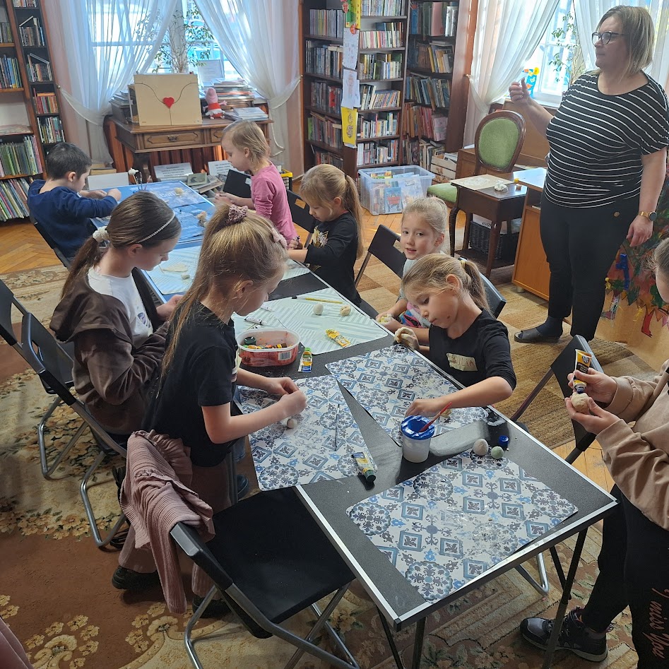 Przy stołach ustawionych wzdłuż sali bibliotecznej siedzą dzieci. Przed dziećmi  leżą kartki, stoją kubeczki z wodą i pędzelkami. Na kartkach częściowo ulepione kształty z gliny i masy solnej.