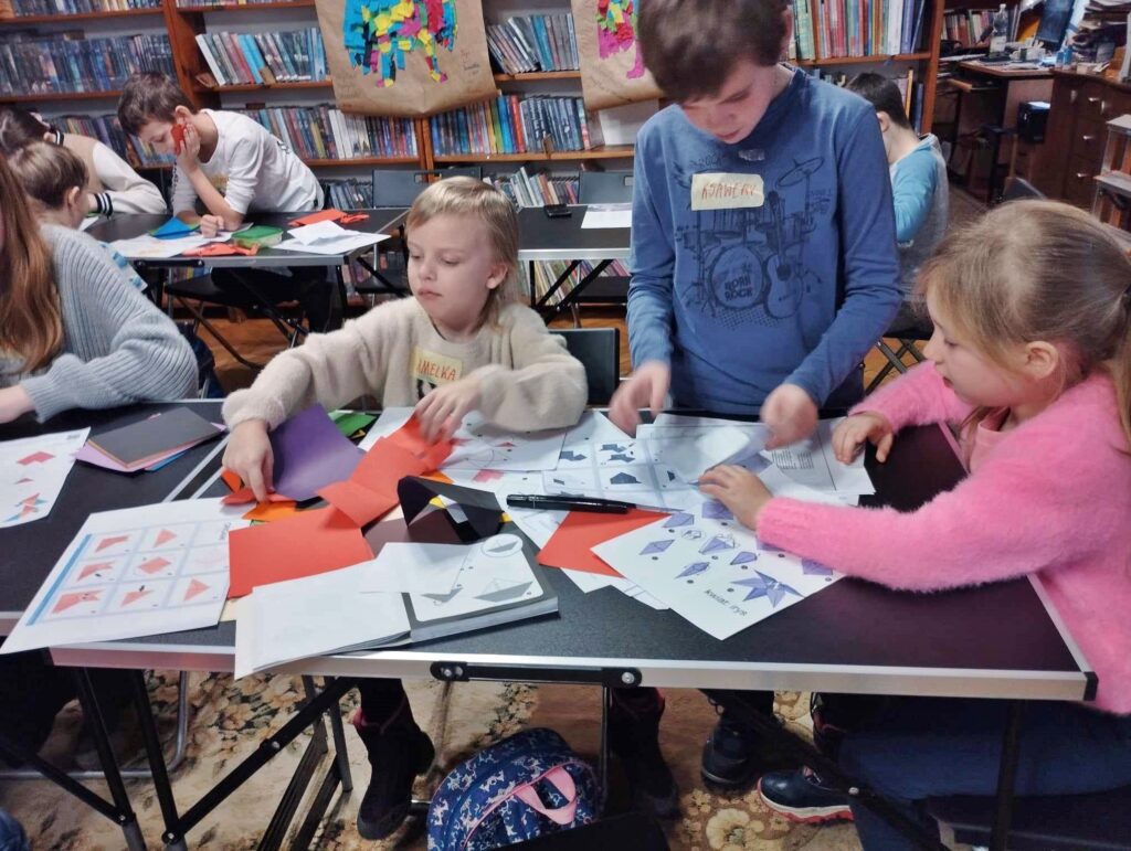 Na stolikach leżą zadrukowane kartki, na których jest instrukcja, jak złożyć obrazek. Przy stolikach siedzą dzieci i próbują składać własne origami.
