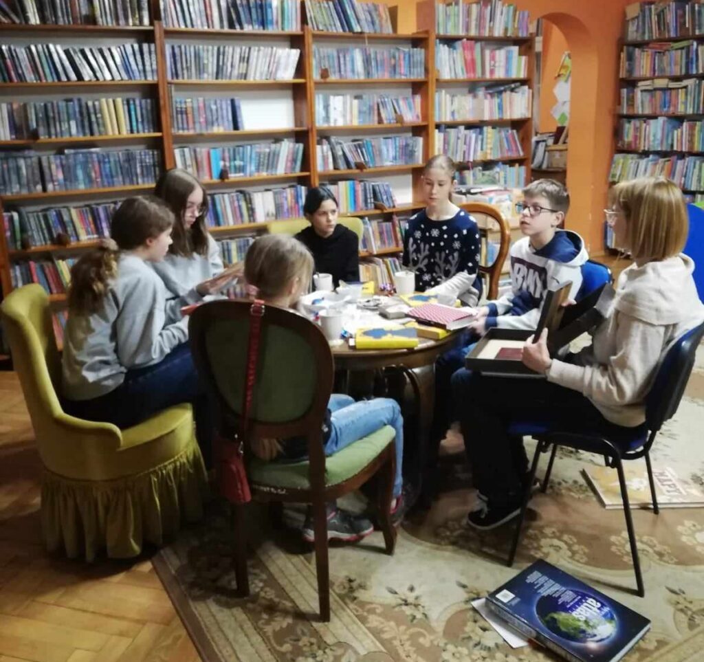 Przy stoliku siedzi 6 osób z Dyskusyjnego Klubu Książki dla dzieci dla klas 5-7 oraz moderatorka. Obok stolika, na dywanie lezy atlas oraz książka o mapach.