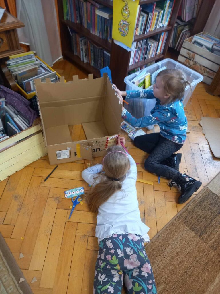 2 dziewczynki. J leży na podłodze i rysuje flamastrem po brązowym kartonie. Druga robi to samo z prawego boku kartonu.