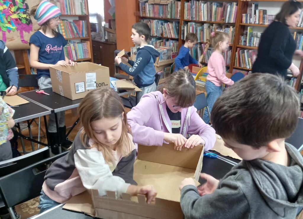 Wzdłuż sali biblioteczne ustawione stoły. Przy stołach dzieci oraz pracownice biblioteki budują zamki i pałace z kartonów i rolek po papierze.