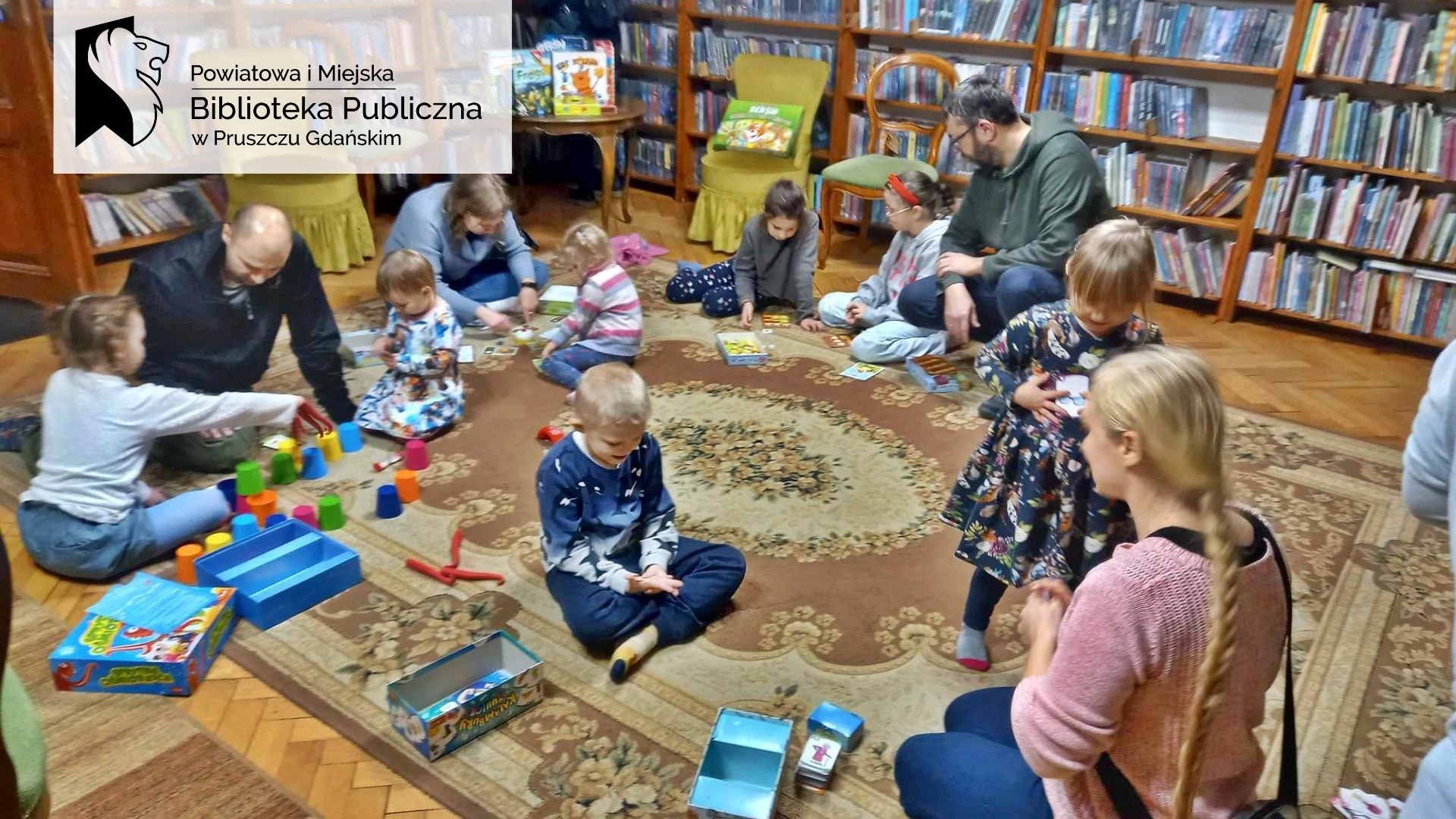 Dorośli i dzieci siedzą na dywanie i grają w planszówki. W tle regały z książkami.