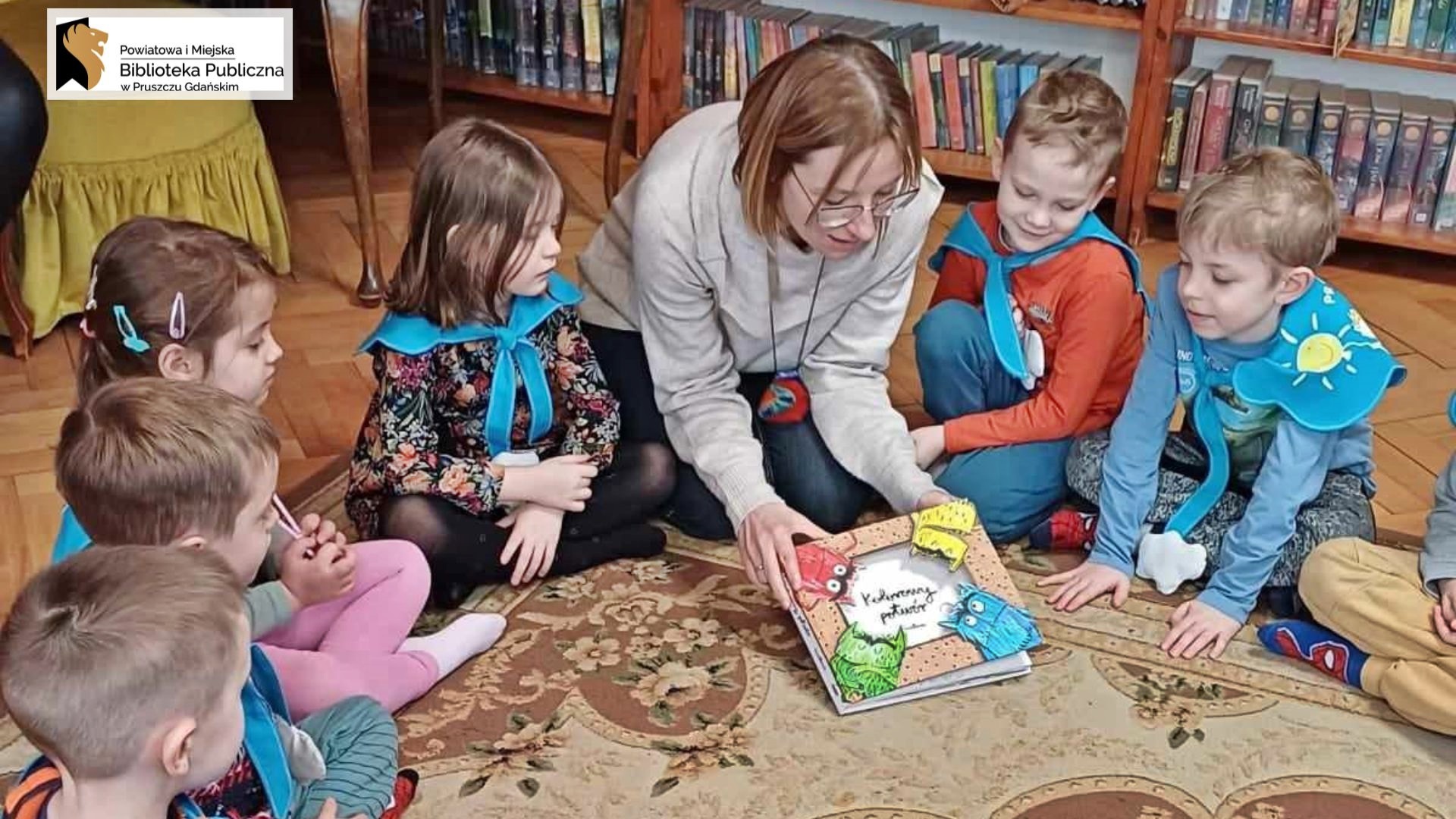 Na dywanie siedzi 5 dzieci i bibliotekarka. Bibliotekarka pokazuje dzieciom książkę pt. Kolorowy potwór.