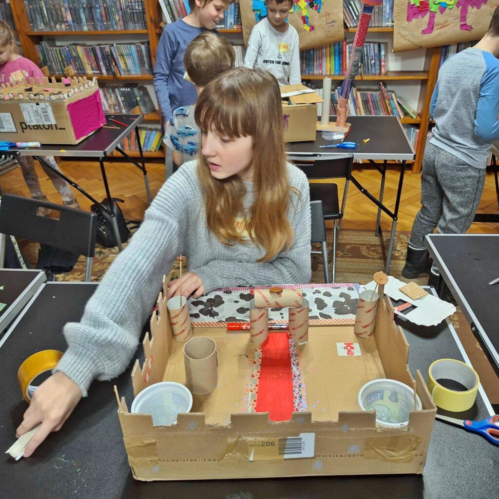 Na pierwszym palnie dziewczynka. Dziewczynka siedzi przy stole. Przed dziewczynką częściowo wykonana makieta zamku.