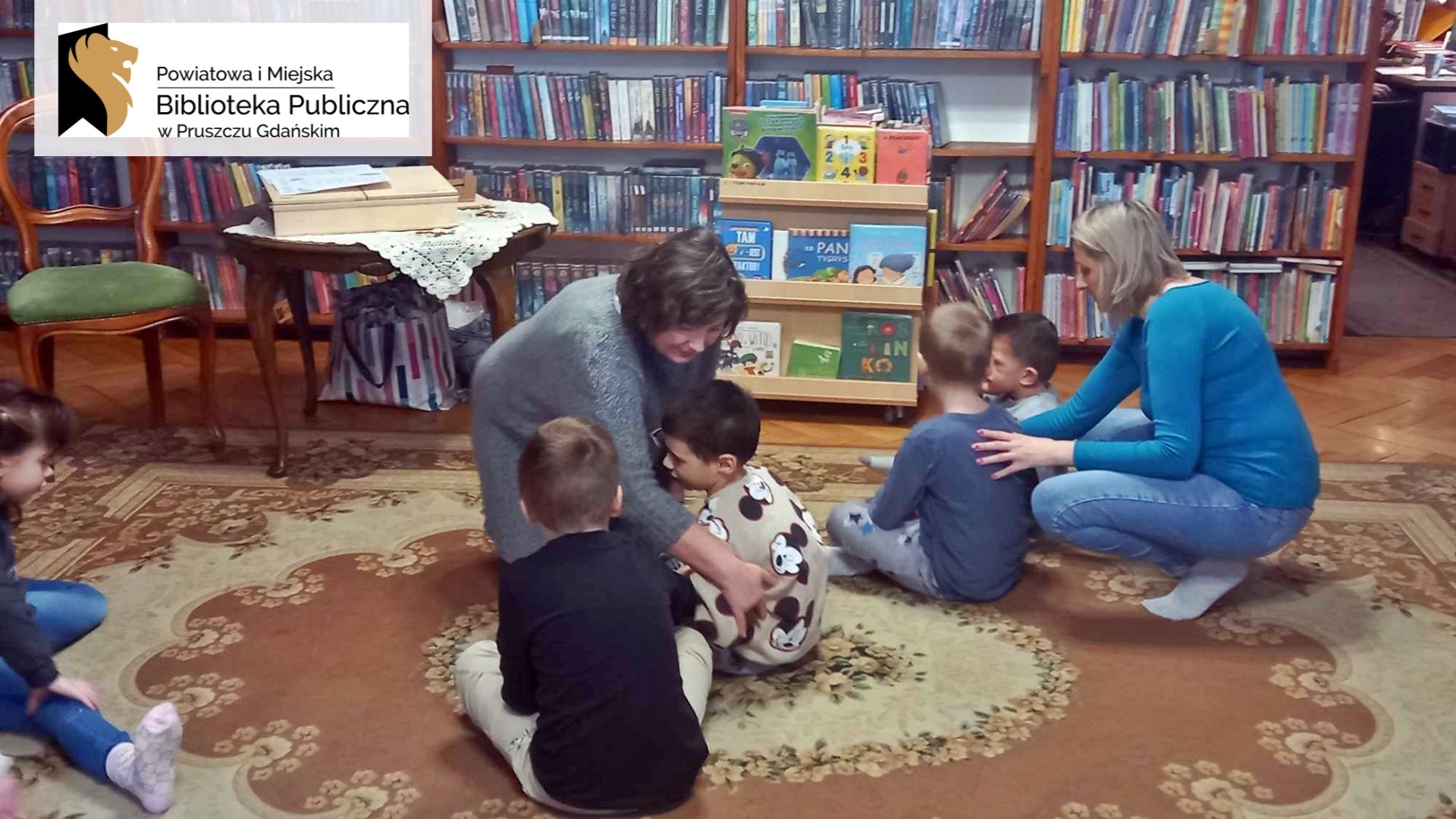 Na dywanie siedzi 4 dzieci. Obok 2 dzieci klęka nauczycielka i robi ręką masażyk pleców. Obok 2 następnych klęka bibliotekarka i również robi masażyk.