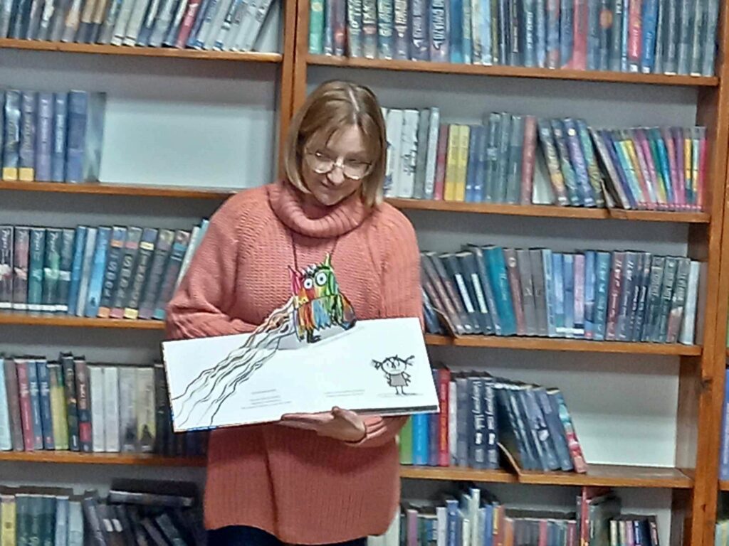 Bibliotekarka trzyma w rękach otwartą książkę. Z książki wystaje przestrzenna ilustracja pokazująca kolorowego potworka. W tle regały z książkami.