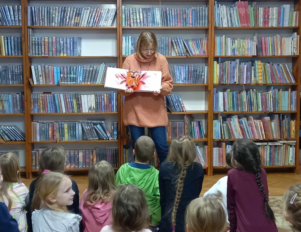 Grupa dzieci siedzi na dywanie i patrzy na bibliotekarkę, która pokazuje książkę. W książce ilustracja kolorowego potworka.