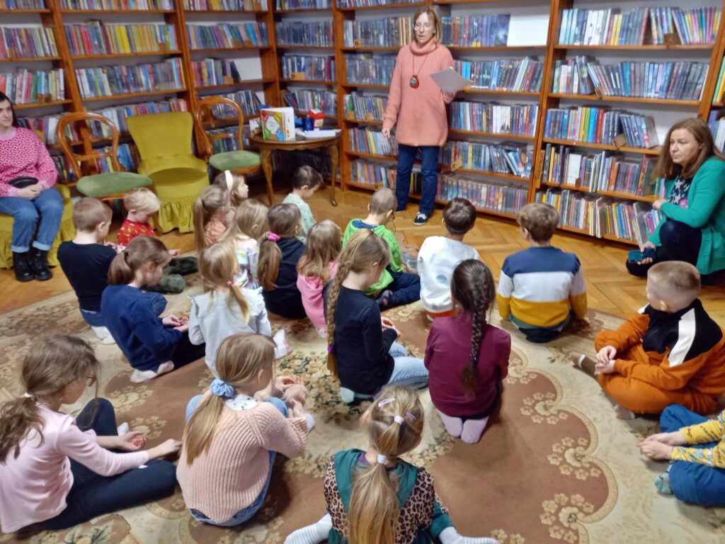 Grupa dzieci siedzi na dywanie i patrzy na bibliotekarkę, która trzyma w ręce kartkę i coś mówi. Obok bibliotekarki stoi stolik, na którym stoi książka, leża kleje i nożyczki.