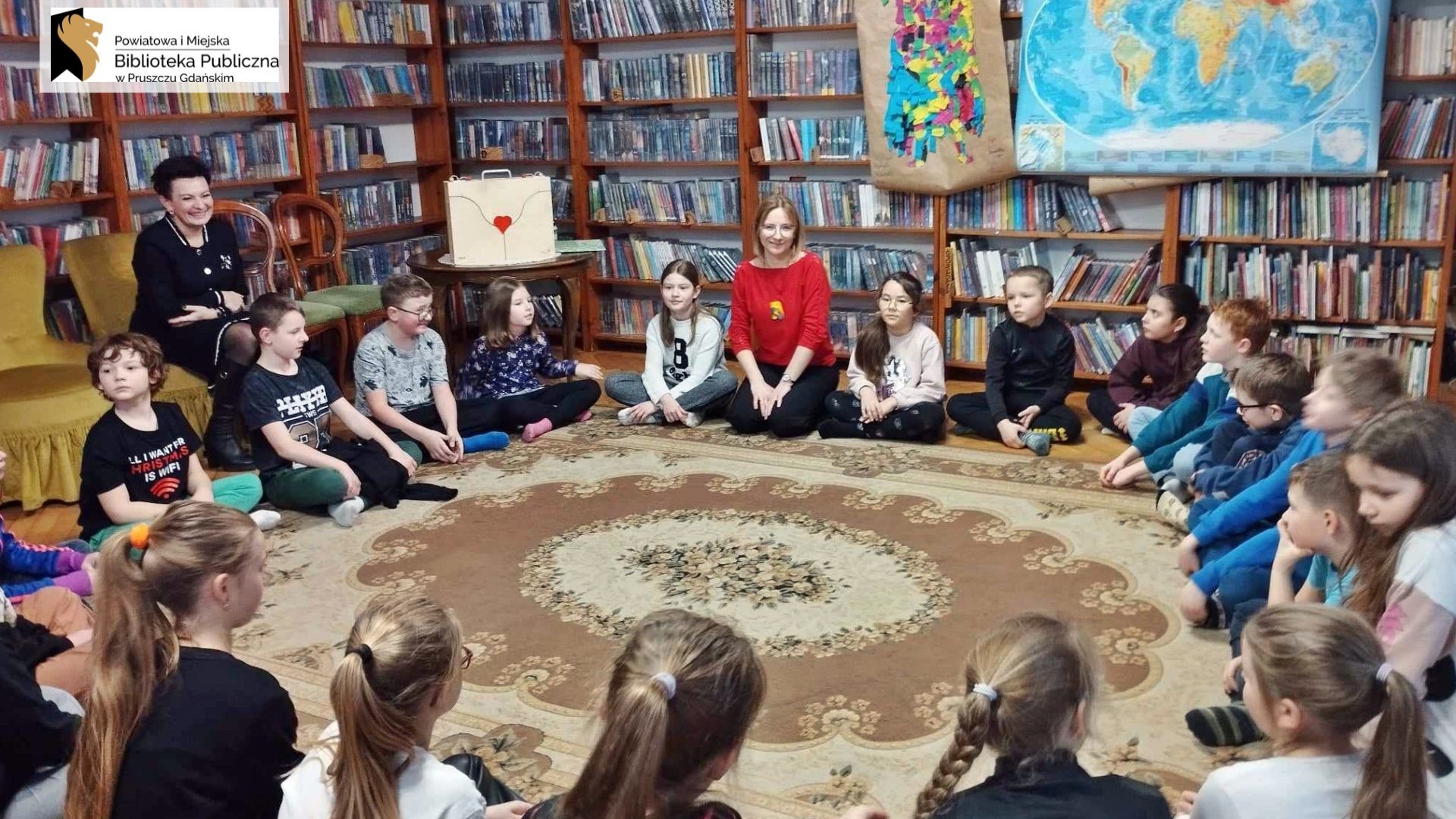 Grupa dzieci siedzi w okręgu na dywanie. Między nimi siedzi bibliotekarka. W tle regały z książkami oraz stolik, na którym stoi drewniany teatrzyk.