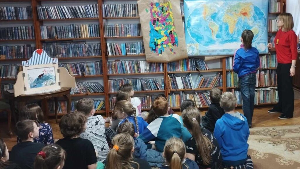 Duża grupa dzieci siedzi na dywanie. Dzieci patrzą przed siebie. Przed nimi stoi chłopiec i bibliotekarka, którzy patrzą na mapę wiszącą na regale z książkami.
