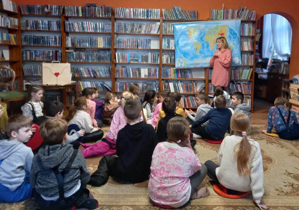 Duża grupa dzieci siedzi na dywanie. Dzieci patrzą w stronę bibliotekarki, która coś mówi. Za bibliotekarką, na regałach z książkami, wisi mapa świata.