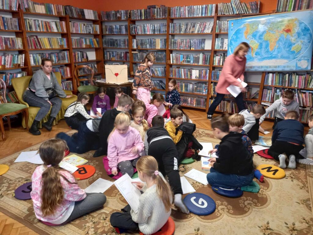 Duża grupa dzieci siedzi na dywanie. Dzieci piszą na kartkach. Na regałach z książkami, wisi mapa świata.