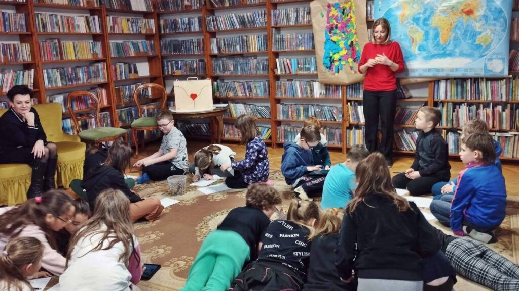 Duża grupa dzieci siedzi i lezy na dywanie. Dzieci piszą coś na kartkach. Na dzieci patrzy bibliotekarka.  W tle regały z książkami. Na 1 z regałów wisi mapa.