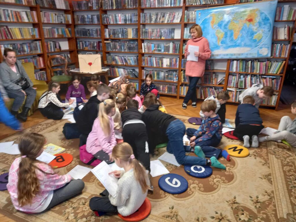Duża grupa dzieci siedzi na dywanie. Przed dziećmi leżą kartki. Dzieci patrzą w stronę bibliotekarki, która trzyma w rękach biała kartkę i coś mówi. Za bibliotekarką, na regałach z książkami, wisi mapa świata.