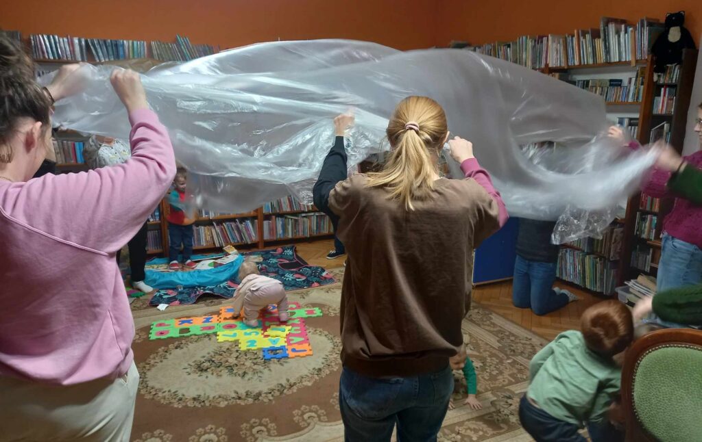 Rodzice i bibliotekarka potrząsają folią malarską. Pod folią, na dywanie, leżą dzieci.