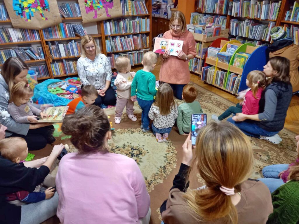 Rodzice i malutkie dzieci. Niektóre dzieci stoją, inne są na rękach u rodziców. Patrzą na bibliotekarkę, która pokazuje ilustrację z czytanej książki. Na obrazku Kicia Kocia oraz: czapka, szalik, rękawiczki i buty.