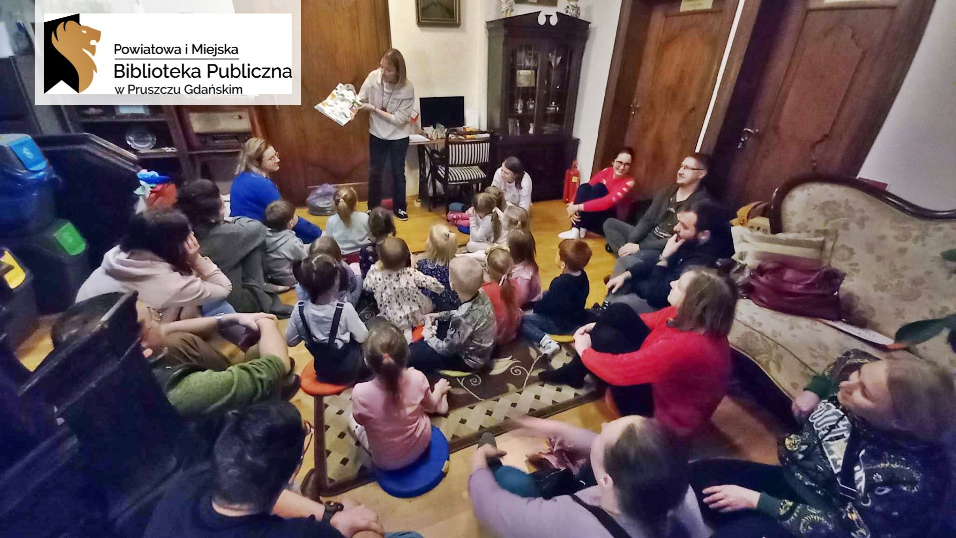 Duża grupa dzieci oraz dorosłych siedzi na dywanie i podłodze. Wszyscy patrzą w stronę bibliotekarki, która pokazuje ilustrację z czytanej książki. Na ilustracji kolorowego, kudłatego stworka.