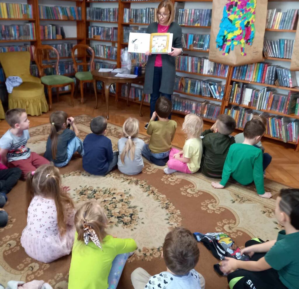 Grupa dzieci siedzi na dywanie i patrzy na bibliotekarkę, która pokazuje książkę. W książce ilustracja kolorowego potworka. Za bibliotekarką regały z książkami. Z prawej strony, na regale zawieszona praca innych dzieci przedstawiająca kolorowego potwora.  