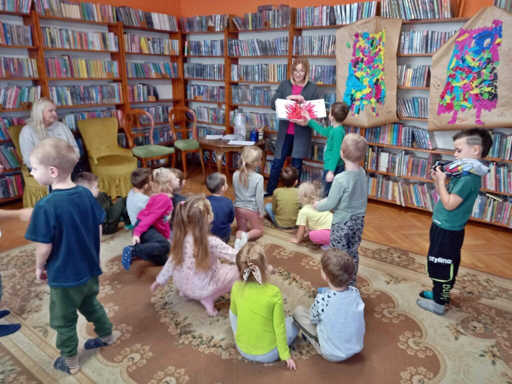 Grupa dzieci siedzi na dywanie i patrzy na bibliotekarkę, która pokazuje książkę. W książce ilustracja kolorowego potworka. Obok stoi chłopiec i wskazuje palcem na ilustrację. Za bibliotekarką regały z książkami. Z prawej strony, na regale zawieszona praca innych dzieci przedstawiająca kolorowego potwora.  