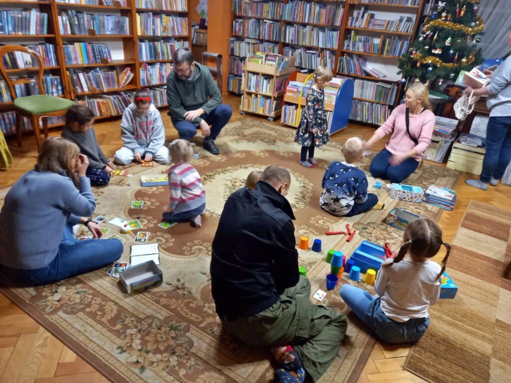 Na dywanie siedzą rodzice i dzieci. Grają w planszówki.