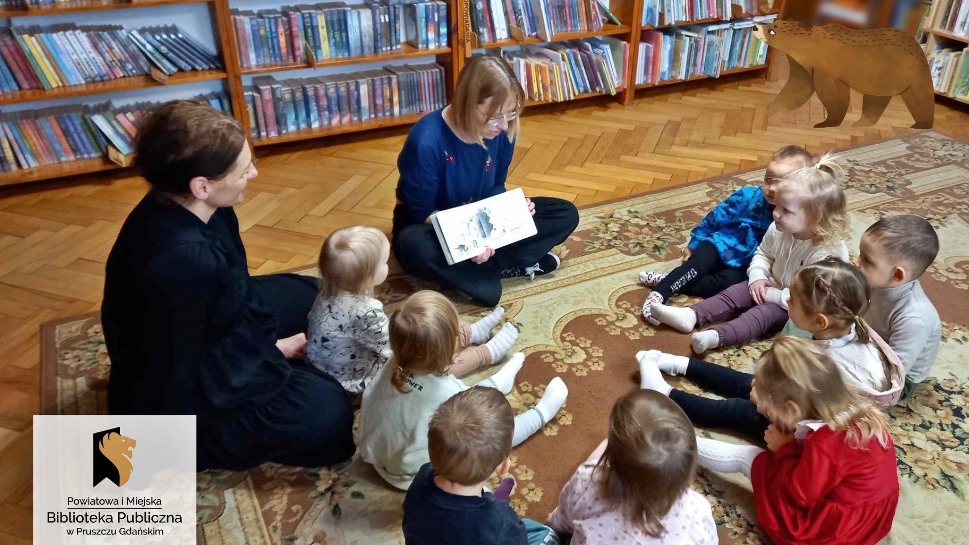 W kółku siedzą malutkie dzieci i nauczycielka. Między dziećmi siedzi bibliotekarka, która czyta książkę i pokazuje ilustracje.