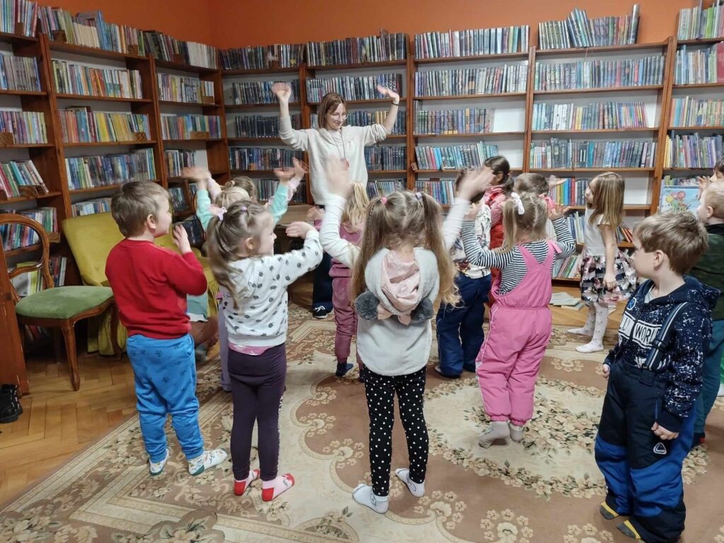 Bibliotekarka stoi przed grupą dzieci i podnosi ręce do góry. Dzieci naśladują bibliotekarkę. 
