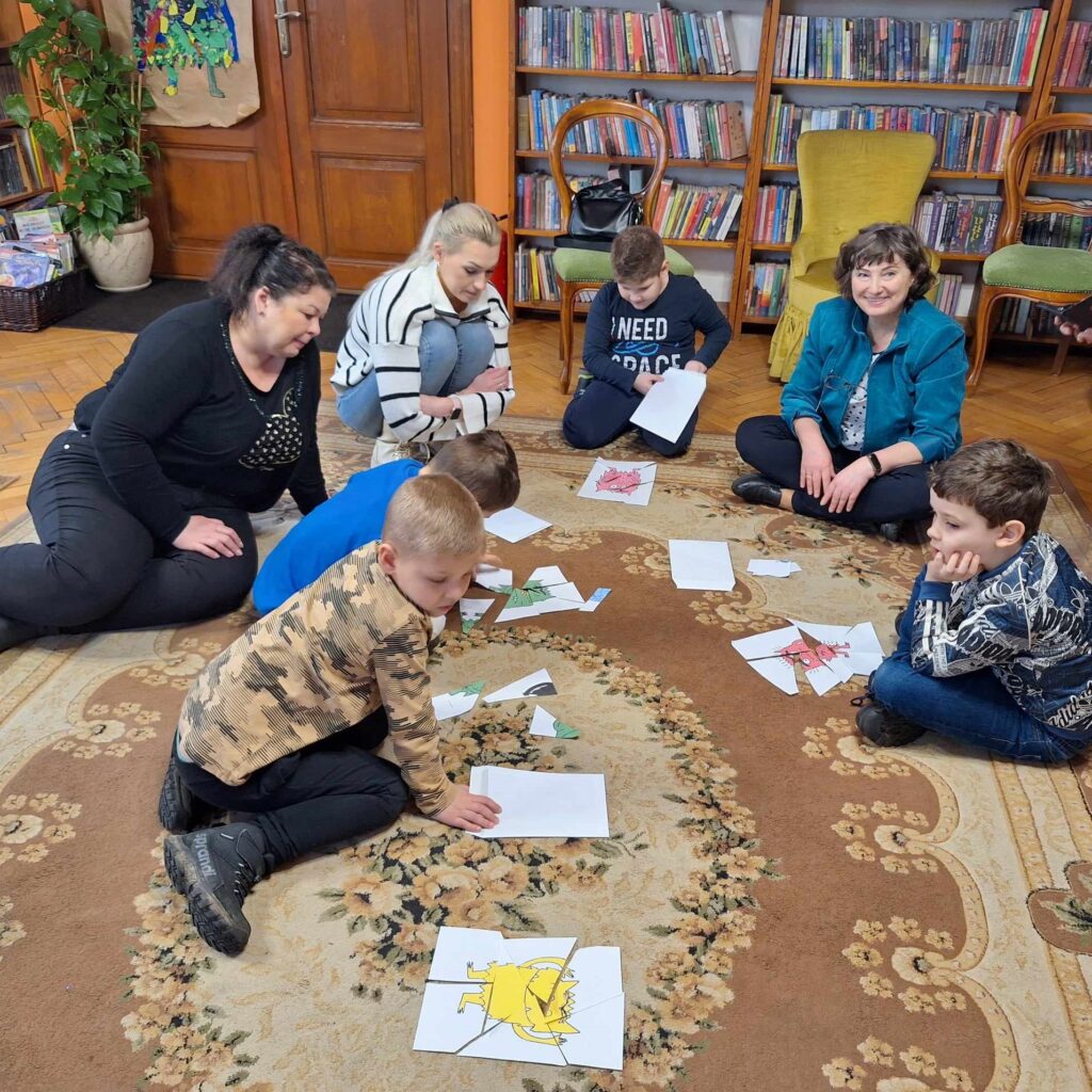 Na podłodze siedzą dzieci. Przed dzieci leży pocięta na puzzle kartka. Dzieci układają obrazek. Obok dzieci siedzi bibliotekarka i 2 opiekunki.
