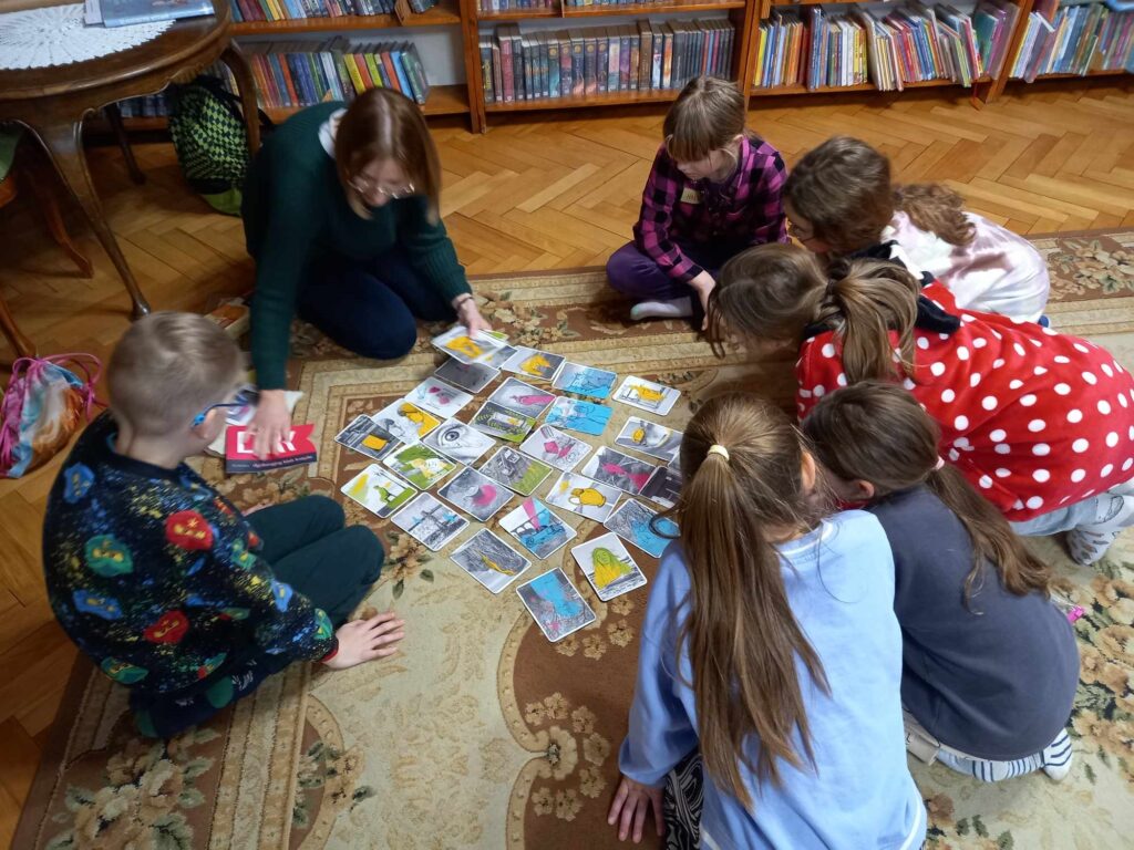 Grupa dzieci i bibliotekarka siedzą na dywanie. Przed nimi rozłożone na dywanie kolorowe karty.