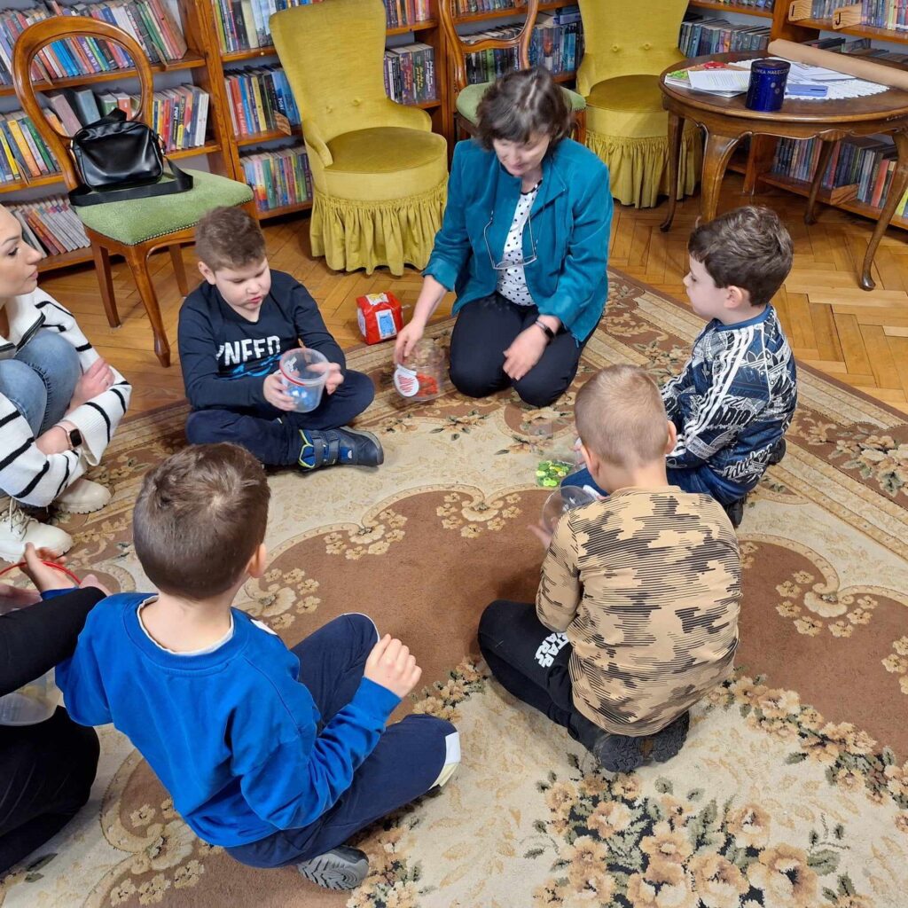 4 dzieci i bibliotekarka siedzi na dywanie. Przed dziećmi stoją 4 plastikowe pudełka oraz leżą na dywanie kolorowe guziki. Dzieci wkładają guziki wg. kolorów do  pudełek.
