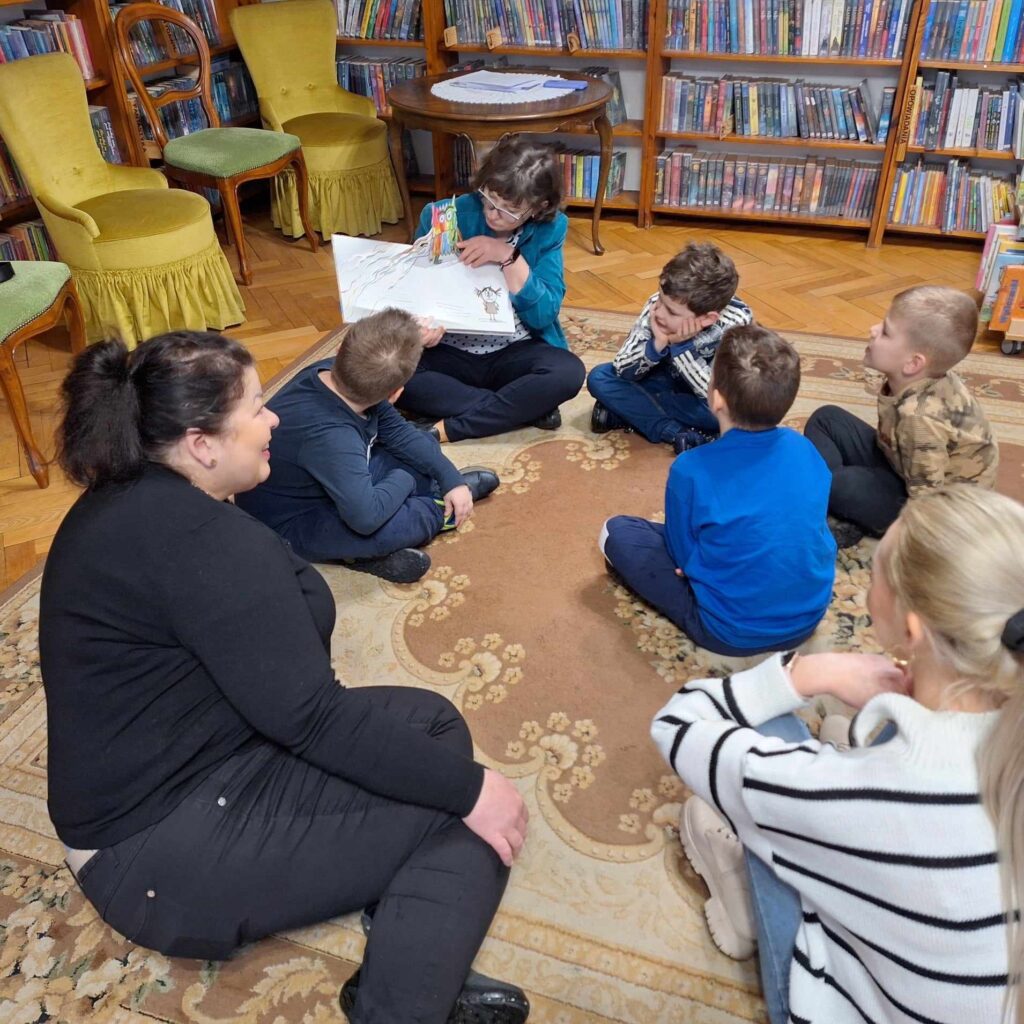 4 dzieci siedzi na dywanie w bibliotece. Dzieci patrzą na bibliotekarkę, która czyta książkę i pokazuje ilustracje. Ilustracja jest przestrzenna i wystaje z niej postać kolorowego potwora.
