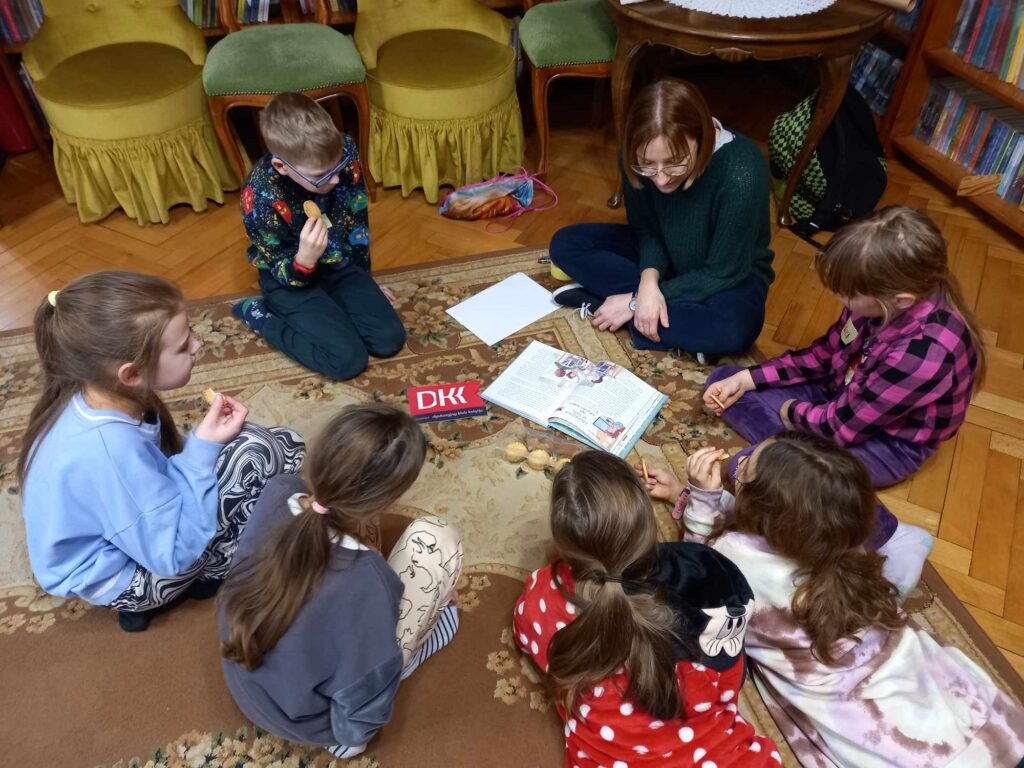 Grupa dzieci na dywanie. Jedzą ciastka. Na dywanie przed nimi leży otwarta książka.