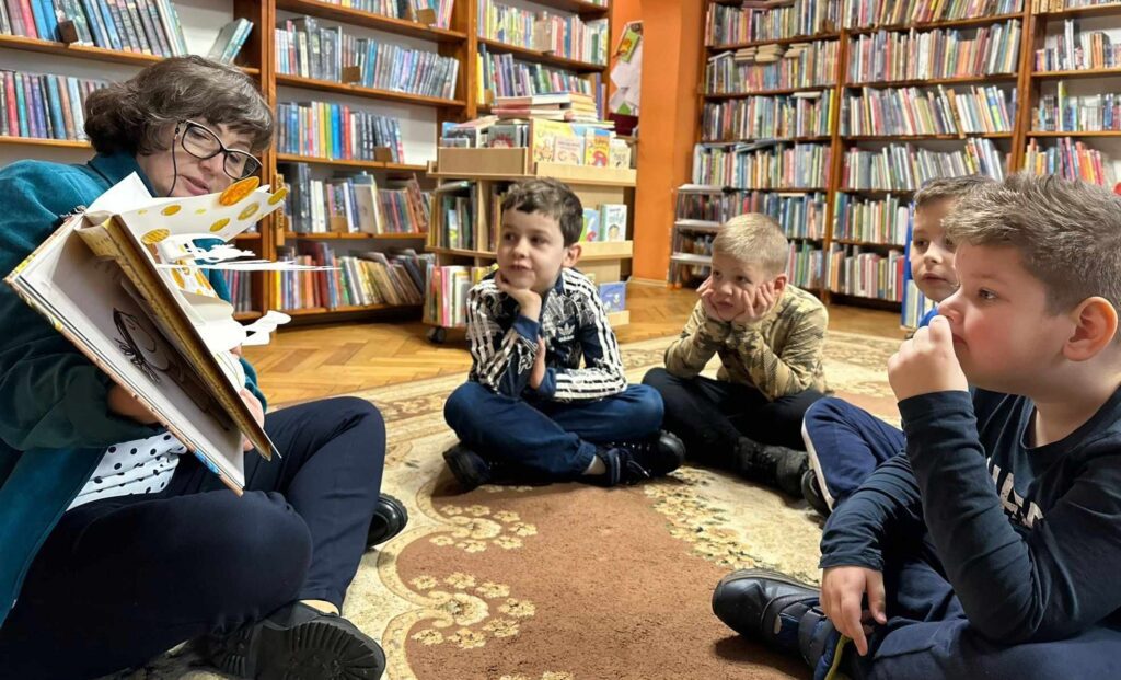 4 dzieci siedzi na dywanie w bibliotece. Dzieci patrzą na bibliotekarkę, która czyta książkę i pokazuje ilustracje. Ilustracja jest przestrzenna i wystaje z niej ilustracja.
