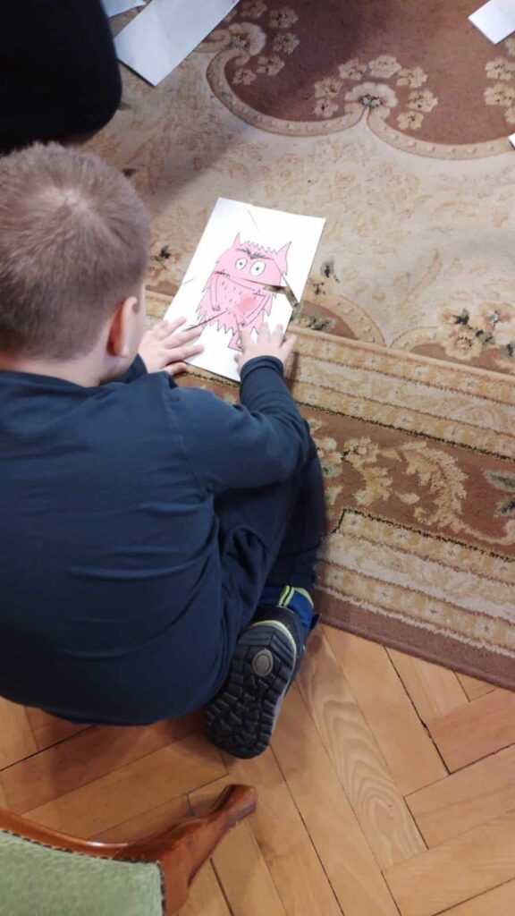 Chłopiec klęka na dywanie i układa z fragmentów kartki różowego potwora. Kartka pocięta jest na puzzle.  

