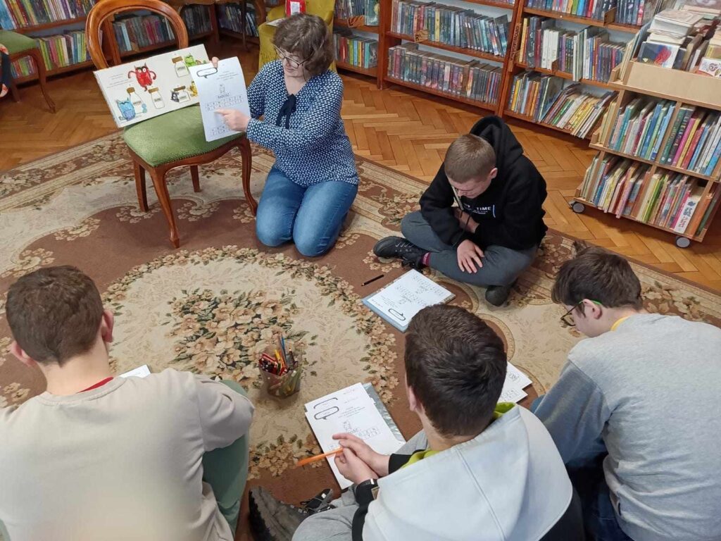 Na dywanie siedzi czworo dzieci. Bibliotekarka pokazuje kartkę i objaśnia im zadania. Obok krzesło z otwartą książką. W tle regały z książkami.