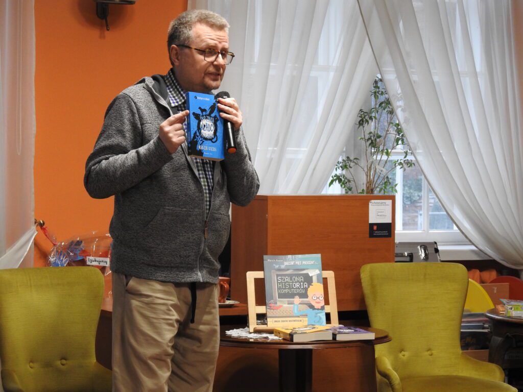 Marcin Kozioł stoi, trzyma w ręku mikrofon i książkę. Na stoliku obok trzy pozycje jego autorstwa.