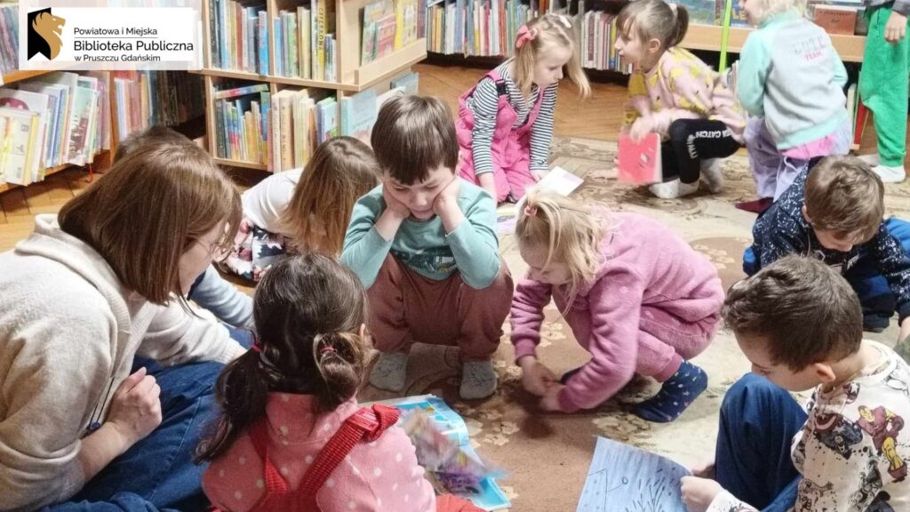 Dzieci siedzą, kucają na dywanie i oglądają książeczki. W tle regały z książkami.