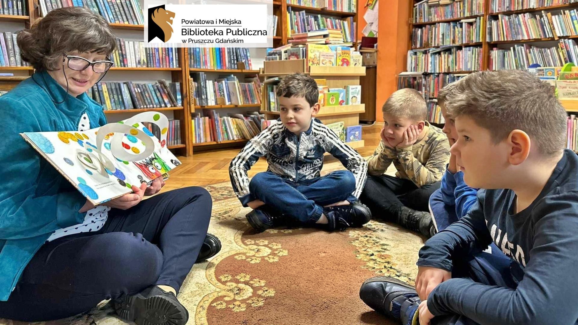4 dzieci siedzi na dywanie w bibliotece. Dzieci patrzą na bibliotekarkę, która czyta książkę i pokazuje ilustracje. Ilustracja jest przestrzenna i wystaje z niej biały w kolorowe kropki zawijas.